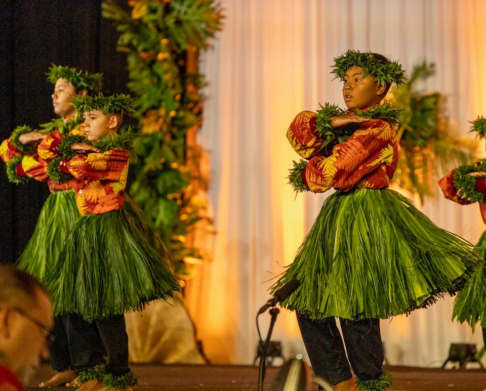 Keiki dancing in green skirts, orange tops, and leafy lei.